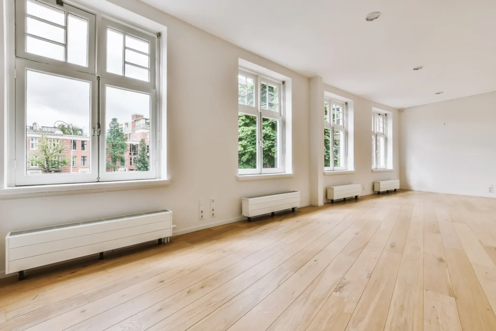 A bright, spacious room showcasing newly installed light-colored hardwood flooring. Large windows provide ample natural light, highlighting the clean lines and quality of the flooring.