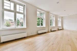 A bright, spacious room showcasing newly installed light-colored hardwood flooring. Large windows provide ample natural light, highlighting the clean lines and quality of the flooring.