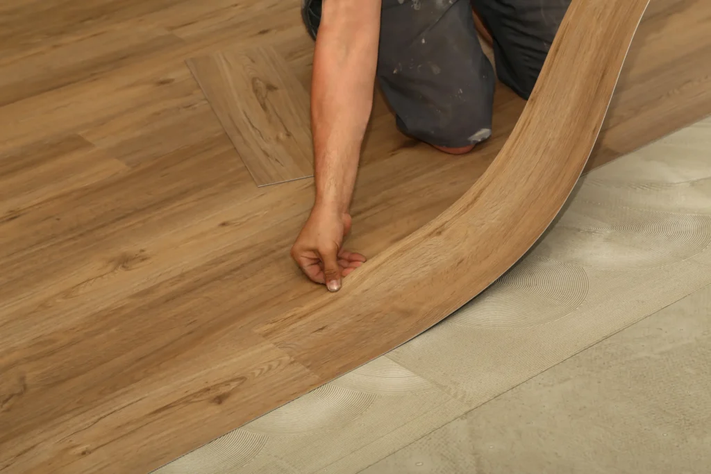 A person's hand carefully placing a luxury vinyl plank during installation, showcasing the precise and detailed nature of the process. The image highlights the modern and stylish appearance of the flooring.